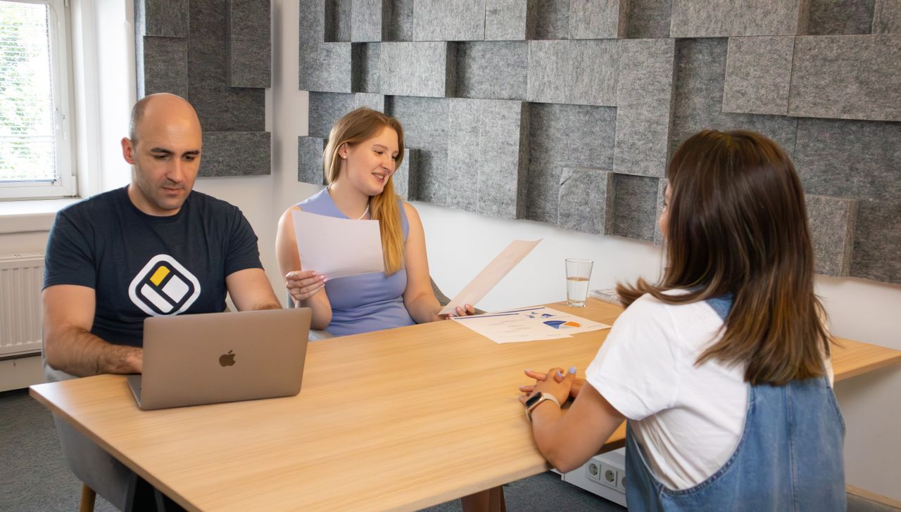 Three colleagues in a modern office discussing work with laptops and documents.
